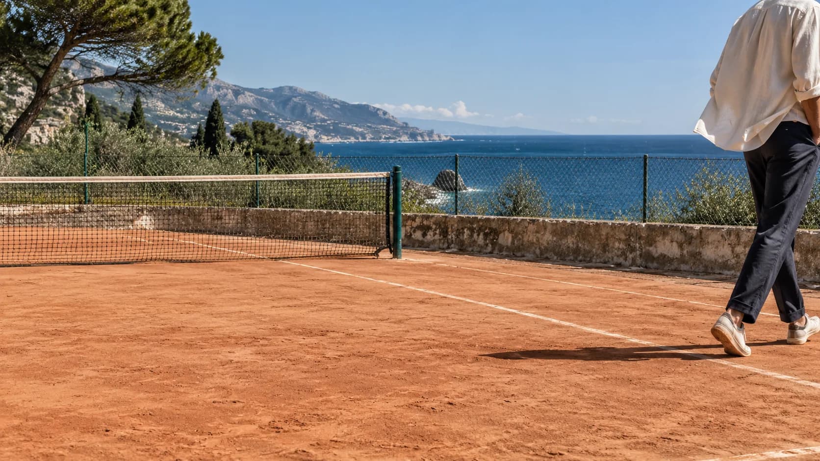 A figure leaving a coastal clay court in bright daylight, with a moving ivory sleeve and blue sea beyond.